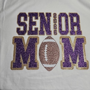 May include: White t-shirt with the words "SENIOR MOM 2023" in purple glitter lettering outlined in gold glitter. A football with brown and white glitter is centered below the text. The shirt is likely for a football mom.