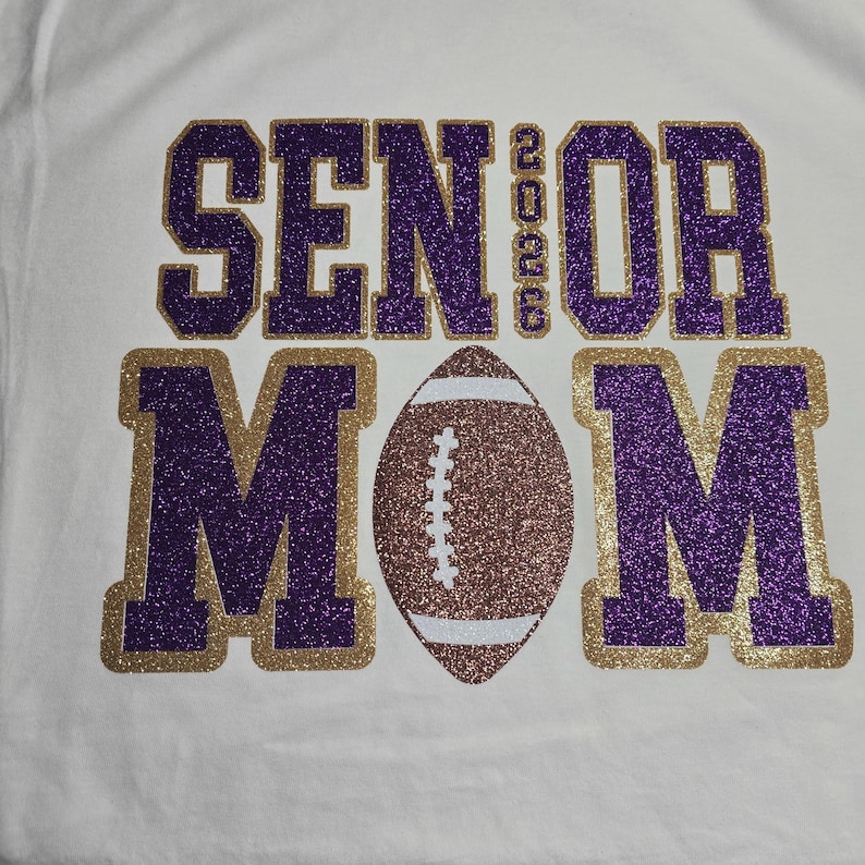 May include: White t-shirt with the words "SENIOR MOM 2023" in purple glitter lettering outlined in gold glitter. A football with brown and white glitter is centered below the text. The shirt is likely for a football mom.