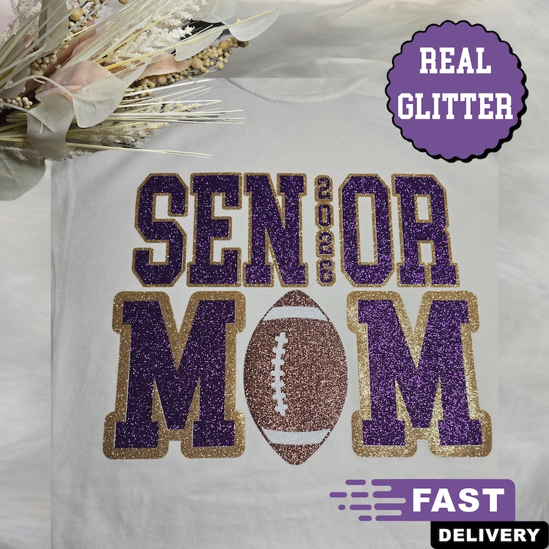 May include: White t-shirt with the words "SENIOR MOM 2026" in purple glitter lettering with gold trim. A glitter football is in the center. A purple circle with "REAL GLITTER" is in the upper right. The words "FAST DELIVERY" are at the bottom.
