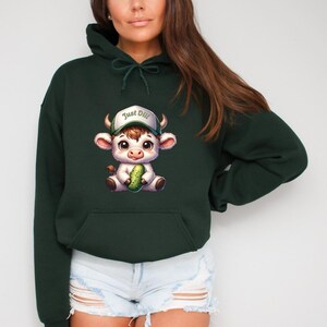 May include: A dark green hooded sweatshirt with a cartoon illustration of a white cow wearing a green baseball cap with the text "Just Dill" on it. The cow is holding a green pickle.