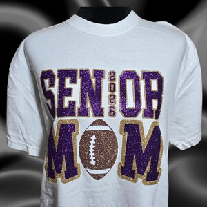 May include: White t-shirt with the words "SENIOR 2023 MOM" in purple and gold glitter lettering. A brown and white football graphic is centered below the text. The shirt has short sleeves and a crew neck.