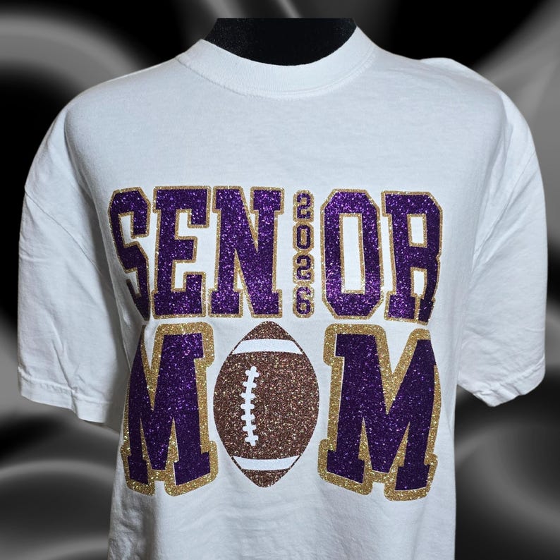 May include: White t-shirt with the words "SENIOR 2023 MOM" in purple and gold glitter lettering. A brown and white football graphic is centered below the text. The shirt has short sleeves and a crew neck.