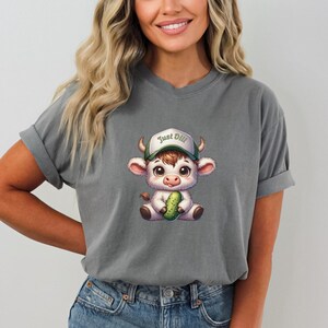 May include: Gray t-shirt with a cartoon illustration of a white cow wearing a green baseball cap that says "Just Dill" and holding a green pickle.
