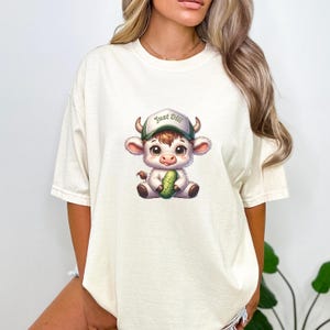 May include: A white t-shirt with a cartoon illustration of a white cow wearing a green and white baseball cap that says "Just Dill" and holding a green pickle.