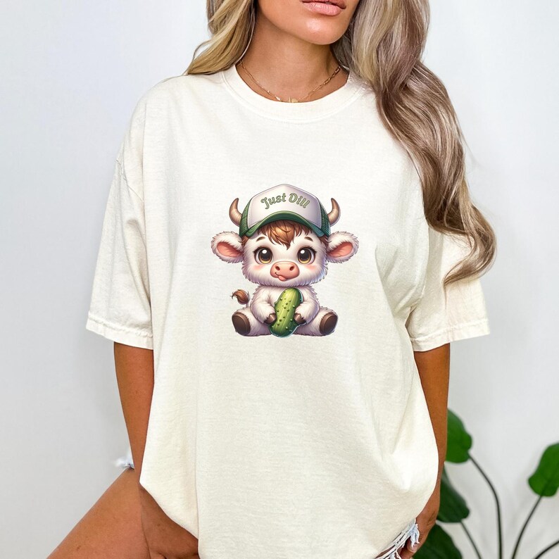 May include: A white t-shirt with a cartoon illustration of a white cow wearing a green and white baseball cap that says "Just Dill" and holding a green pickle.