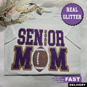 May include: White t-shirt with purple glitter text that reads "SENIOR 2026 MOM" and a brown football graphic. The text is outlined in gold glitter. A purple circle with "REAL GLITTER" is in the upper right corner.