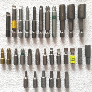 May include: A selection of metal screwdriver bits, including flathead, Phillips head, and Torx types. The bits vary in size and shape, with some featuring colour-coded markings for easy identification. The tools are arranged on a white surface.