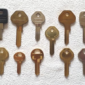 Könnte beinhalten: Elf Schlüssel in verschiedenen Formen und Größen auf einer weißen, strukturierten Oberfläche. Die Schlüssel sind hauptsächlich gold- und silberfarben, einige haben schwarze Kunststoffelemente. Auf einem Schlüssel ist das Wort "Master" zu sehen.