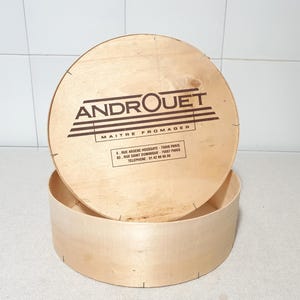 Könnte beinhalten: Eine runde Holzkiste mit Deckel. Der Deckel trägt den Text "ANDROUET MAITRE FROMAGER" und die Kontaktdaten des Unternehmens.