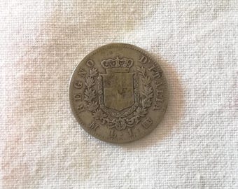 May include: A silver coin from the Regno D'Italia era, featuring a crowned shield surrounded by ornate detailing and the inscription "REGNO D'ITALIA". The coin is displayed on a textured, off-white surface.