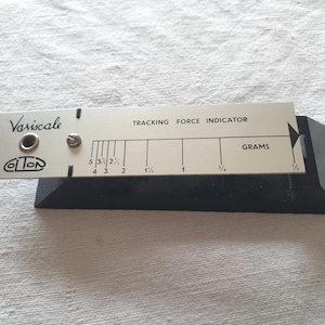 Op de afbeelding: Een Variscale naalddrukmeter met een zilverkleurige metalen schaal op een zwarte voet. De schaal bevat zwarte tekst met de vermelding Variscale, Tracking Force Indicator en Grams, met numerieke markeringen van 1/3 tot 5.