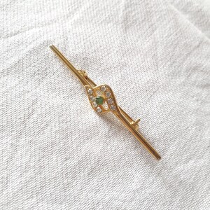 Può includere: Un fermaglio d'oro con una pietra verde circondata da strass trasparenti. Il fermaglio ha la forma di una chiave stilizzata.
