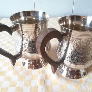 Op de afbeelding: Twee zilverkleurige metalen mokken met donkerbruine houten handvatten. De mokken hebben een bloemmotief in het metaal geëtst en staan op een kleine voet. De handvatten zijn gebogen. De mokken staan op een geel-wit geruit tafelkleed.