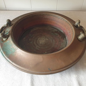 Op de afbeelding: Een grote, ronde koperen schaal met een brede, platte rand en twee messing handgrepen aan weerszijden. Het metalen oppervlak vertoont een verweerde patina met zichtbare groene oxidatievlekken en een donkere, getextureerde bodem.