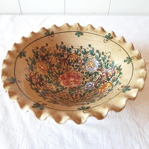 Op de afbeelding: Een beige keramische kom met een geschulpte rand, handbeschilderd met een bloemenmotief. De bloemen zijn in tinten rood, geel, blauw en oranje, met groene bladeren en ranken. De kom heeft een diameter van ongeveer 30 cm.