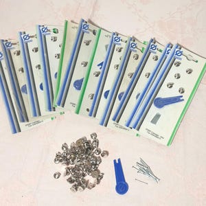 Op de afbeelding: Een assortiment oorbelruggen en accessoires. Verschillende verpakkingen met oorbelruggen zijn gerangschikt met een stapel zilverkleurige oorbelruggen, een blauwe tool en kleine zilveren pinnen. De verpakkingen hebben blauwe, groene en witte accenten.