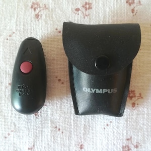 Könnte beinhalten: Schwarze Fernbedienung mit einem roten Knopf und dem Text "REMOTE CONTROL RC-34" auf der Vorderseite. Die Fernbedienung befindet sich in einem schwarzen Lederetui mit dem Wort "OLYMPUS" auf der Vorderseite.