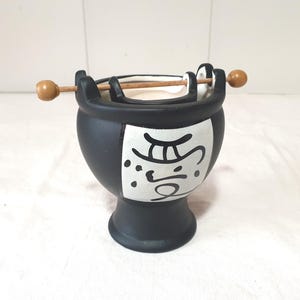 Può includere: Un bruciatore di incenso in ceramica nera con un design rettangolare bianco con calligrafia in stile asiatico. Il bruciatore ha una base a piedistallo e un manico con un bastoncino di legno e due palline di legno. Il bruciatore di incenso è progettato per contenere incenso.