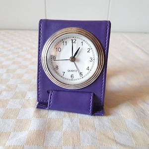 May include: A purple leather clock stand with a white analogue clock face that reads "QUARTZ".