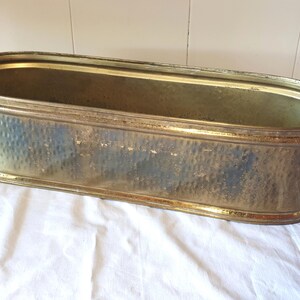 Pode incluir: Uma jardineira de formato oval feita de latão com uma textura martelada na superfície externa. O metal apresenta uma pátina envelhecida e desgastada com manchas escuras visíveis e um acabamento dourado metálico. O recipiente tem uma borda lisa.