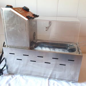 Peut inclure: Une friteuse professionnelle en acier inoxydable avec un corps métallique rectangulaire doté de fentes de ventilation horizontales, un pare-éclaboussures en acrylique transparent, un panneau de commande avec cadran noir et interrupteur orange.