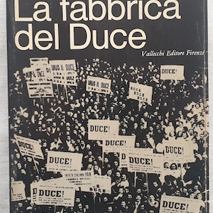May include: Book cover with the title "Dino Biondi La fabbrica del Duce" in white text on a black background. The cover features a black and white photo of a crowd holding signs that say "DUCE!" and "VIVA IL DUCE".