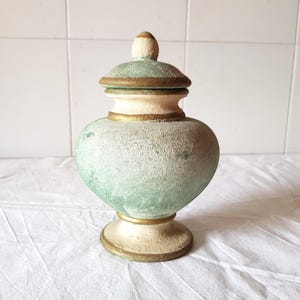 Könnte beinhalten: Eine grüne Keramikvase mit Deckel, verziert mit goldenen Verzierungen und einem weißen Sockel. Die Vase hat eine abgenutzte Oberfläche, die ihr einen Vintage-Look verleiht.