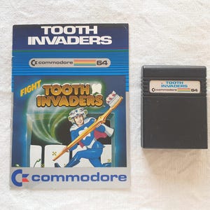 Könnte beinhalten: Ein Tooth Invaders Spielehandbuch für den Commodore 64 mit einem blau gekleideten Superhelden, der eine riesige Zahnbürste hält, neben einer schwarzen rechteckigen Spielkassette mit weißem Etikett, das Tooth Invaders und das Commodore 64 Logo zeigt.