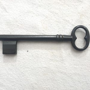 Könnte beinhalten: Ein schwarzer Metallschlüssel mit einem dekorativen, doppellappigen Griff und einem rechteckigen Bart. Der Schlüssel liegt horizontal auf einer weißen Oberfläche. Das Design des Schlüssels deutet auf einen Vintage- oder Antik-Stil hin.