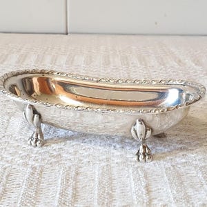 Może przedstawiać: Mała, srebrna miseczka w kształcie wanny. Miseczka ma ozdobną, sznurowatą obwódkę i stoi na trzech nóżkach w kształcie pazurów. Wnętrze miseczki odbija światło.