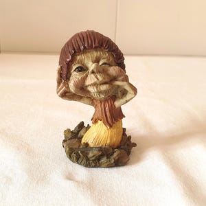 Op de afbeelding: Een grillig beeldje van een lachende gnome met een bruine muts en een gele en bruine outfit. De gnome knipoogt en houdt zijn wangen vast. Het beeldje staat op een voet van bruin en groen textuurmateriaal.
