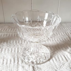 May include: A clear glass pedestal bowl featuring a scalloped rim and a multifaceted diamond-cut pattern across the exterior surface. The bowl is supported by a thick, circular base that also displays a textured, geometric diamond-cut design.