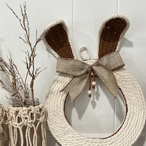 May include: A white rope wreath with brown bunny ears and a burlap bow. The wreath is decorated with small wooden beads.
