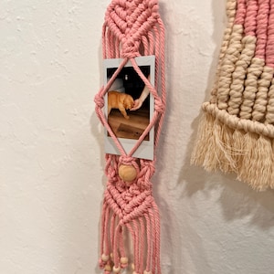 May include: A pink macrame photo holder with a wooden ring at the top and a wooden bead at the bottom. The holder displays a small photo of a cat. Additional macrame wall hanging is visible on the right.