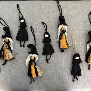 May include: A collection of eight handmade hanging ornaments featuring figures crafted from black, white, and orange yarn. Some figures wear pointed hats and hold small brooms, while others are simple black silhouettes with wooden bead heads.