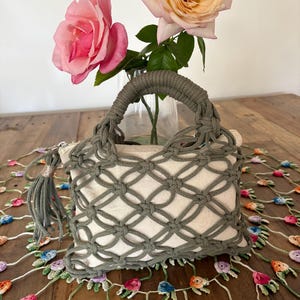 May include: A rectangular beige canvas handbag featuring an olive green macrame overlay with a knotted lattice pattern. The bag includes two matching olive green rope handles and a decorative tassel attached to the silver zipper pull.
