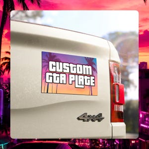May include: A rectangular custom license plate sticker attached to the tailgate of a white truck. The sticker features a sunset gradient background with palm tree silhouettes and the bold white text "CUSTOM GTA PLATE" with a black outline.