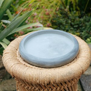 Puede incluir: Una bandeja redonda de hormigón gris claro con un borde elevado descansa sobre un taburete tejido. El taburete está hecho de cuerda y bambú de color natural. El fondo presenta follaje verde y un toque de flores rosas.