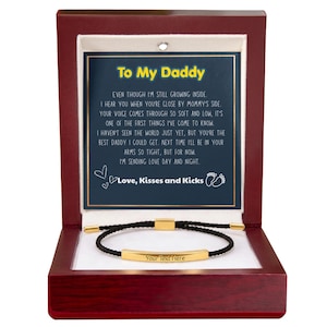 May include: A dark wood gift box containing a black cord bracelet with a gold-tone metal bar engraved with the text Your Text Here. The box lid features a printed message starting with To My Daddy, followed by a poem and the text Love, Kisses and Kicks.