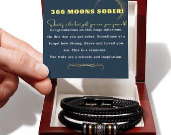 May include: A black braided leather bracelet with a metal clasp engraved with the words love you forever, featuring silver and gold-toned beads, presented in a dark wood box with a card reading 366 MOONS SOBER! and a congratulatory message.