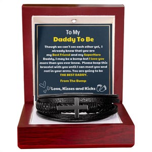 May include: A black leather bracelet with a silver cross, presented in a red wooden box. The box contains a card with the text "To My Daddy To Be" and a message from "The Bump". The bracelet is a thoughtful gift.