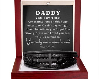 May include: A black leather multi-strand bracelet featuring a central cross charm, presented in a mahogany-coloured wooden box. The interior lid displays the text: DADDY YOU GOT THIS! Congratulations on this huge milestone. On this day you got sober. Sometimes you forget how Strong, Brave and Loved you are. This is a reminder. You truly are a miracle and inspiration.