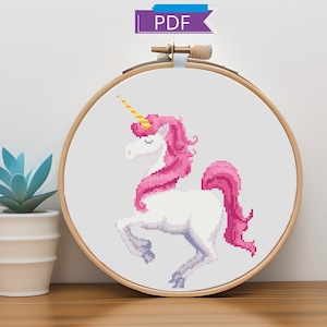 Puede incluir: Un patrón de punto de cruz de un unicornio blanco con una crin y cola rosadas, enmarcado en un aro de madera. El unicornio está de pie sobre sus patas traseras con la cabeza girada hacia un lado.