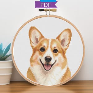 May include: A cross-stitch pattern featuring a portrait of a tan and white Corgi dog with pointed ears and a happy expression, displayed within a round wooden embroidery hoop. A purple tag with the white text PDF is attached to the top.