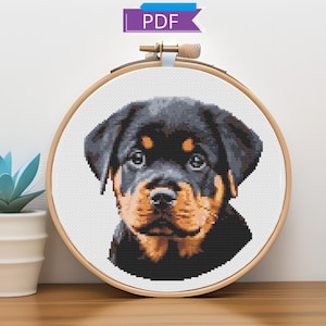 May include: Cross stitch pattern of a Rottweiler puppy in a wooden embroidery hoop. The puppy has black fur with brown markings and is looking directly at the viewer.