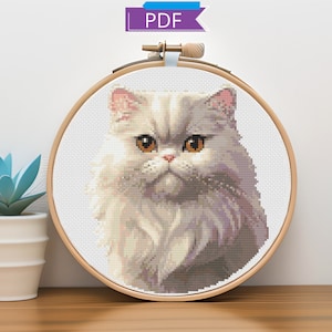 May include: Cross stitch pattern of a white Persian cat with orange eyes in a wooden embroidery hoop. The cat is looking directly at the viewer.