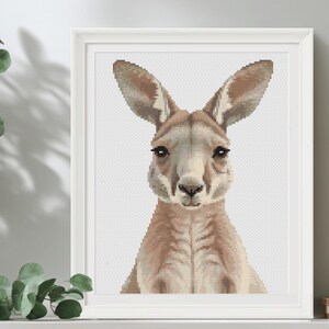 May include: A framed cross-stitch picture of a kangaroo. The kangaroo is depicted with tan and brown fur, large ears, and dark eyes. The artwork is presented in a white frame, ideal for home decoration.