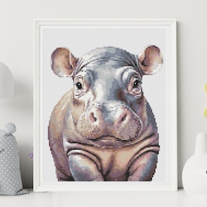 Op de afbeelding: Een close-up van een baby-nijlpaard met een lichtgrijs en bruin kleurenschema. Het nijlpaard heeft een brede, platte neus en kleine, ronde oren. Het nijlpaard kijkt recht in de camera.