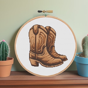 May include: A cross-stitch embroidery hoop with a design of two brown cowboy boots. The boots are stitched in a pixelated style.