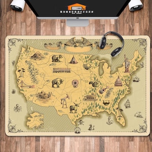 May include: A rectangular desk mat featuring a vintage-style map of the United States with illustrations of landmarks and wildlife. The mat has a cream-colored background with a decorative border and the word "Rushmore" is visible. A pair of black headphones rests on the mat.